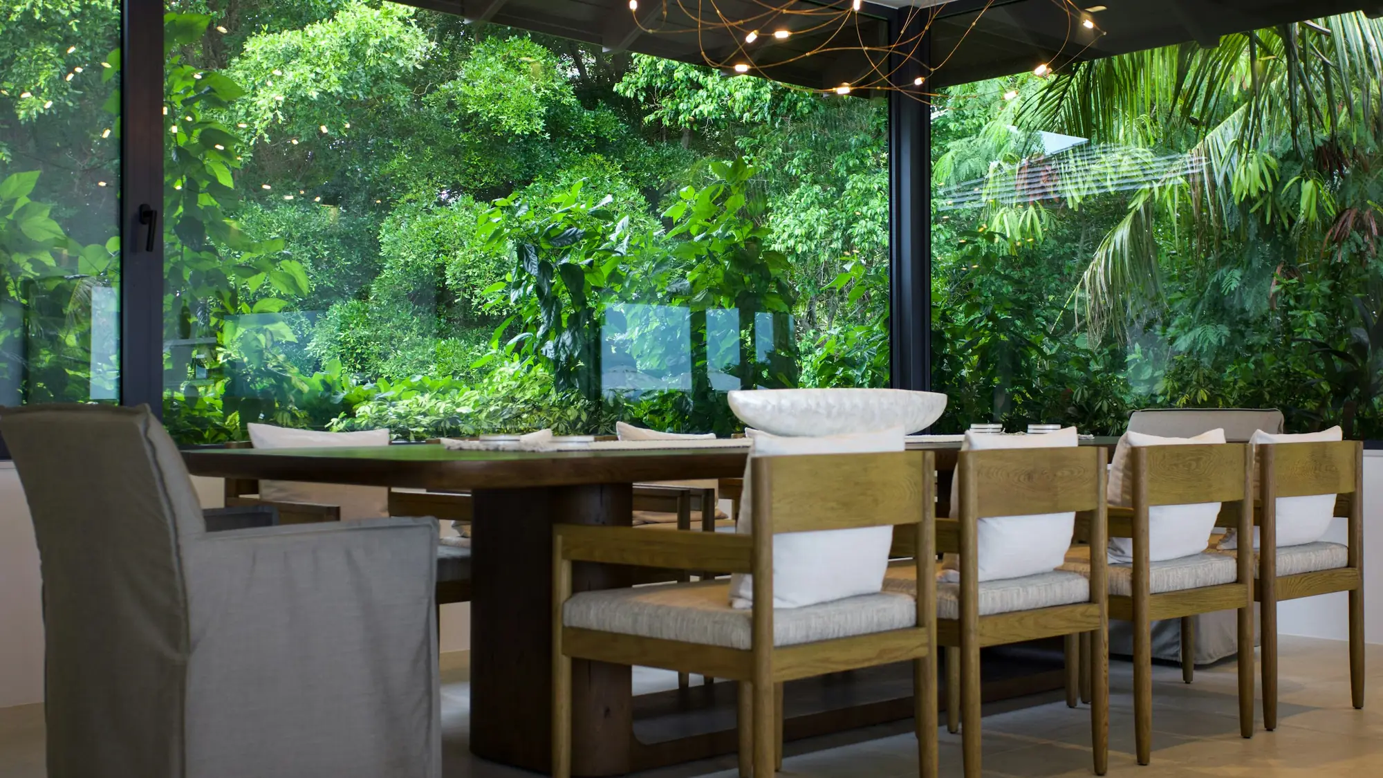 A long timber table in a glass-roofed room overlooking a garden