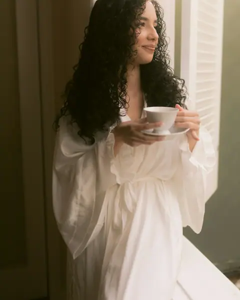 A woman in a white robe holding a cup of tea by the window