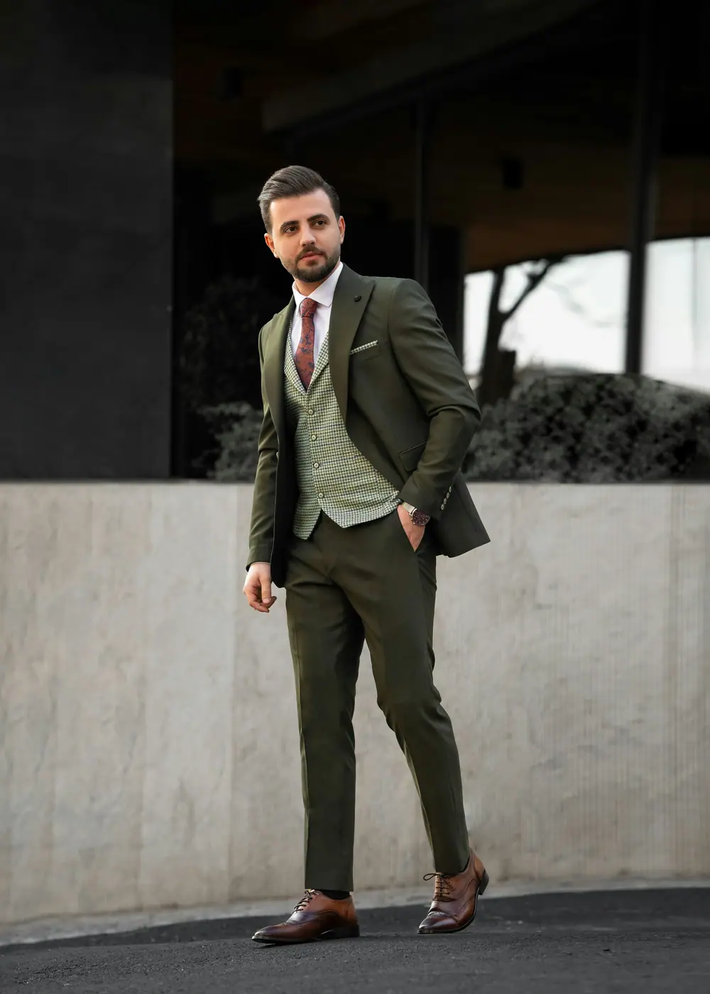 A man in a dark green three-piece suit walking past a concrete wall