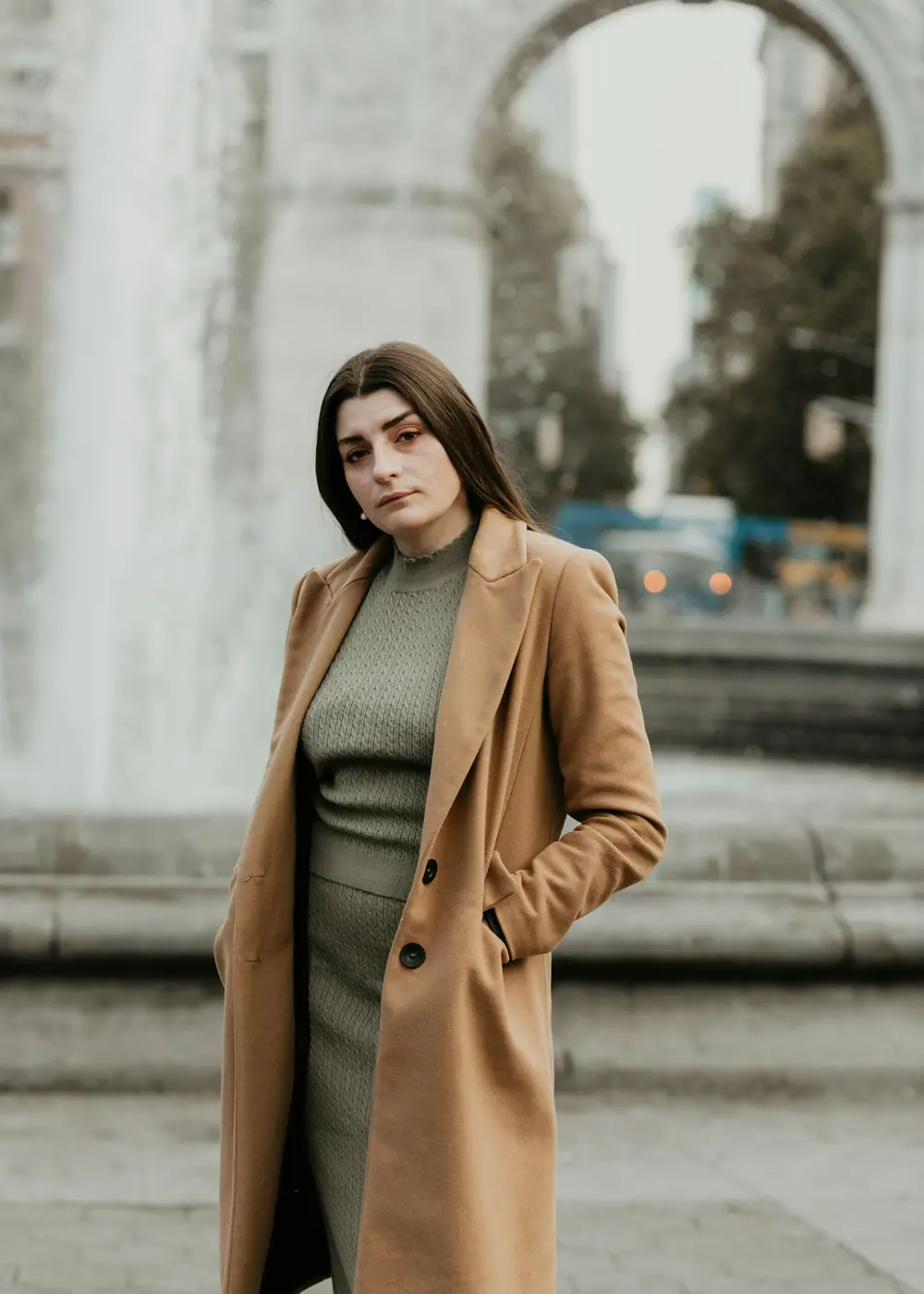 A woman in a long camel coat standing by a fountain